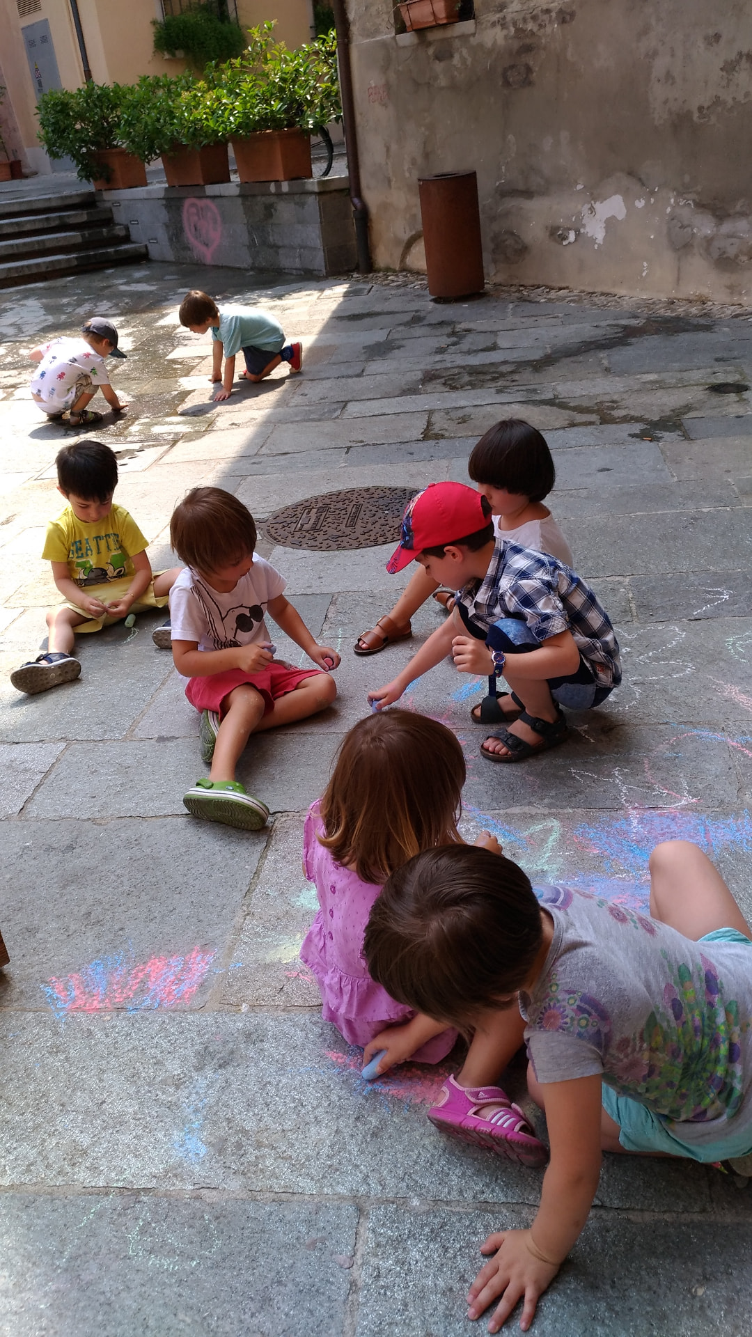 Bambini seduti sul selciato che disegnano con i gessetti in un vicolo ombreggiato