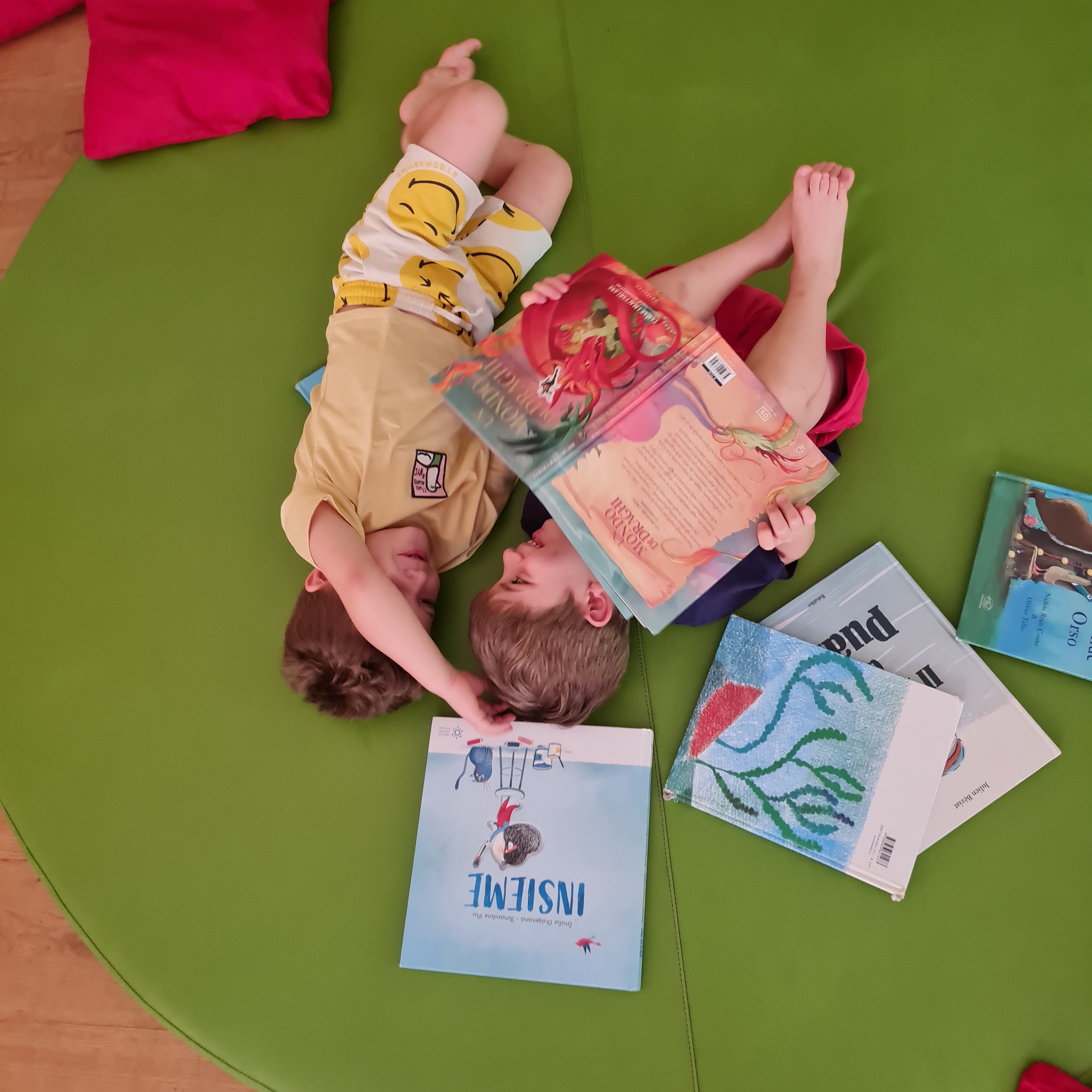 Due bambini sdraiati su un materassino verde che sfogliano insieme libri illustrati