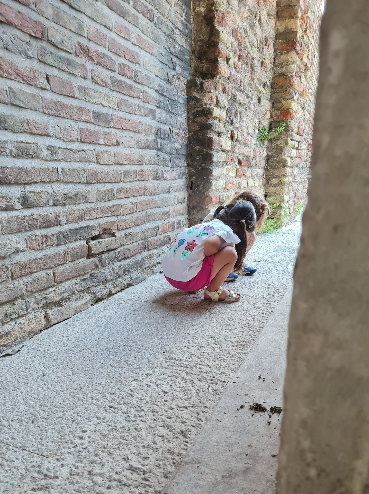 Due bambine accovacciate a terra osservano qualcosa sul pavimento di un passaggio all’aperto, tra pareti di mattoni a vista